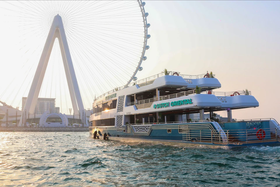 A large double-deck boat labeled "Dutch Oriental" on water near a ferris wheel at sunset.