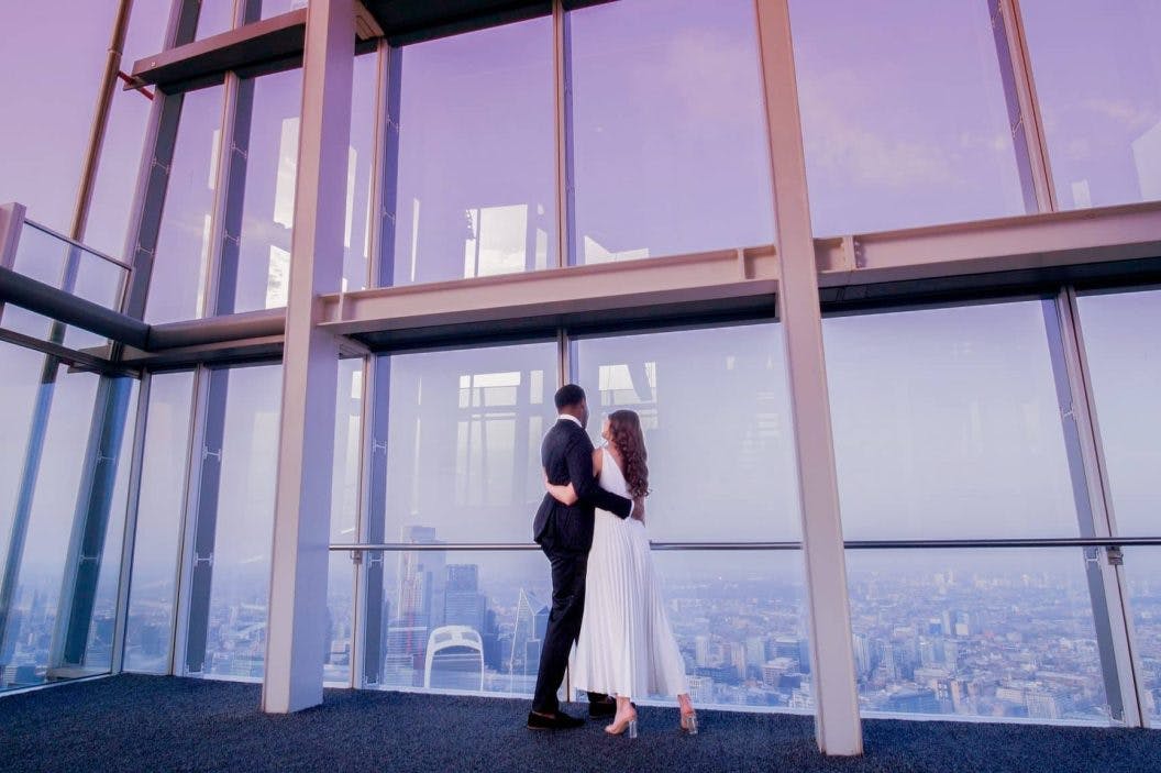 Para w stroju formalnym obejmuje się, patrząc na duże okno z widokiem na panoramę miasta, pod sufitem oświetlonym fioletem.