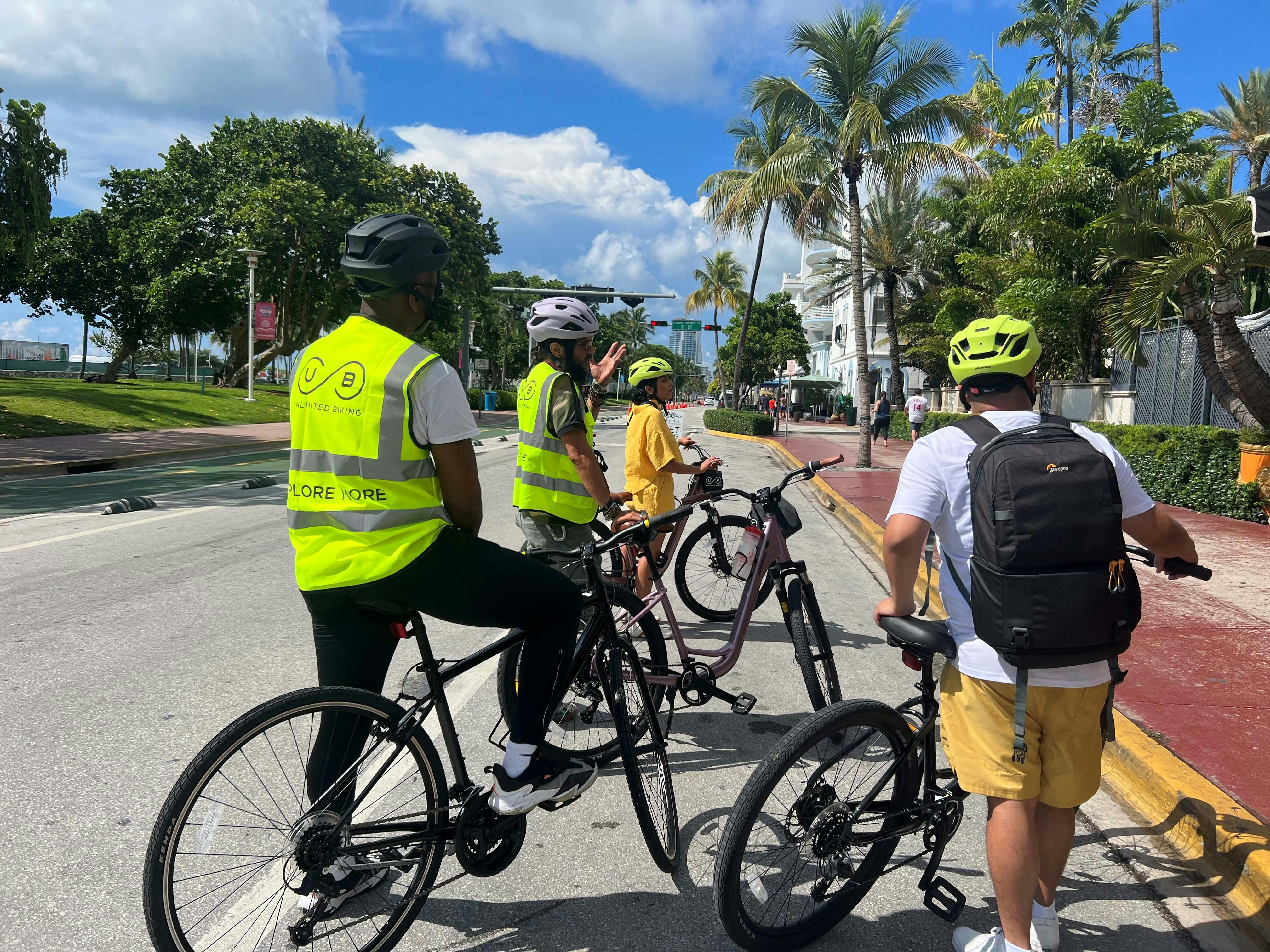 Skupina cyklistů v helmách a bezpečnostních vestách se zastaví na ulici lemované palmami pod částečně zataženou oblohou.