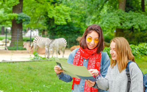 Dos mujeres con ropa informal mirando un mapa en un parque al aire libre con cebras visibles al fondo.