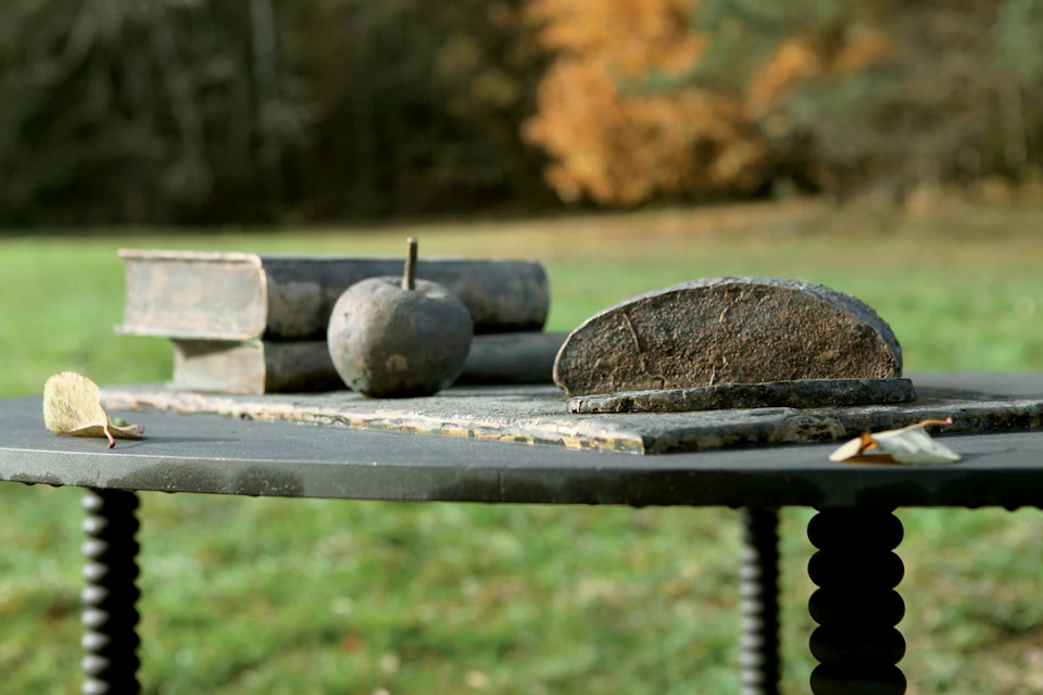 Outdoor table with old books, a round stone, and a broken grindstone, set against an autumn background with trees and grass.
