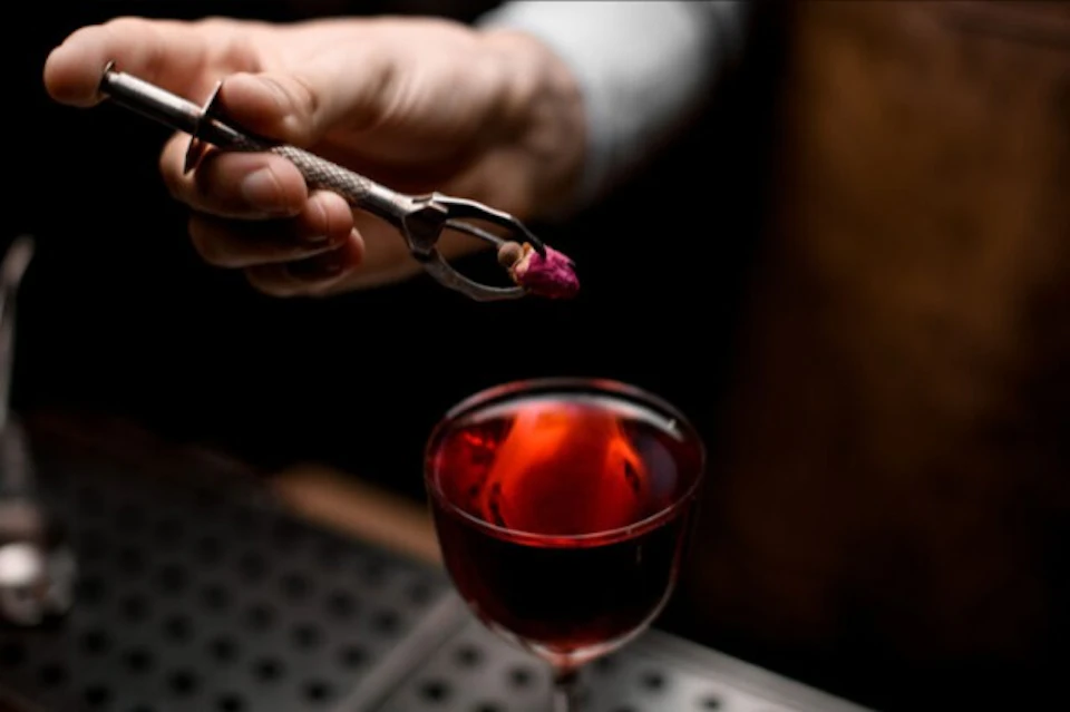 A hand holding tweezers places a small flower bud into a glass of red liquid.