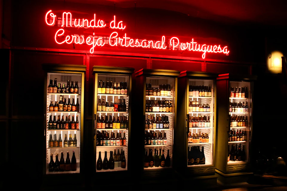 A display of refrigerators filled with bottles of Portuguese craft beer under a neon sign reading, 'O Mundo da Cerveja Artesanal Portuguesa.'