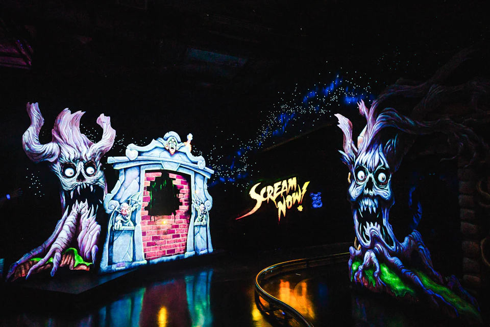 A dark ride scene with a spooky theme, featuring eerie artwork, glowing decorations, and the text 'Scream Now!' lit up.