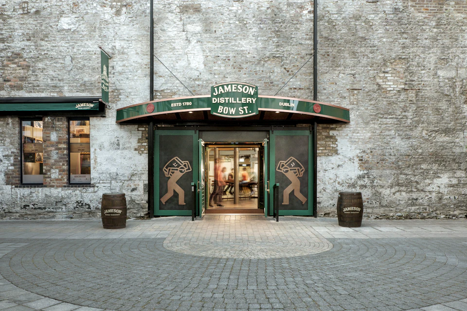 Entrance of Jameson Distillery Bow St. with green doors, whiskey barrel decor, and people visible inside.