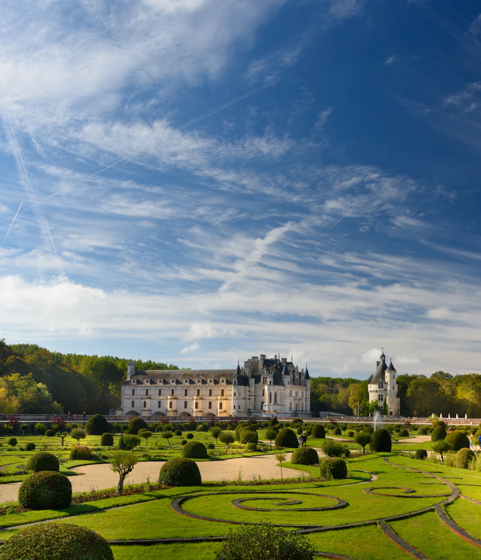Ornate château with manicured gardens and neatly clipped hedges under a blue sky with scattered clouds and contrails.