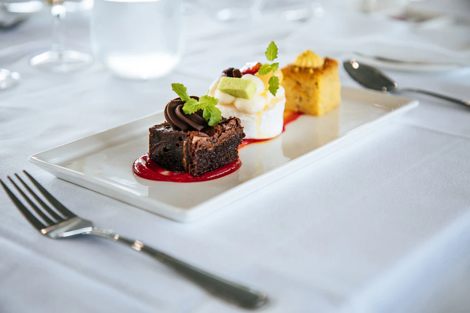 A dessert platter featuring a brownie with chocolate garnish, a fluffy cake with fruit and cream, and a yellow cake with icing on a white tablecloth.