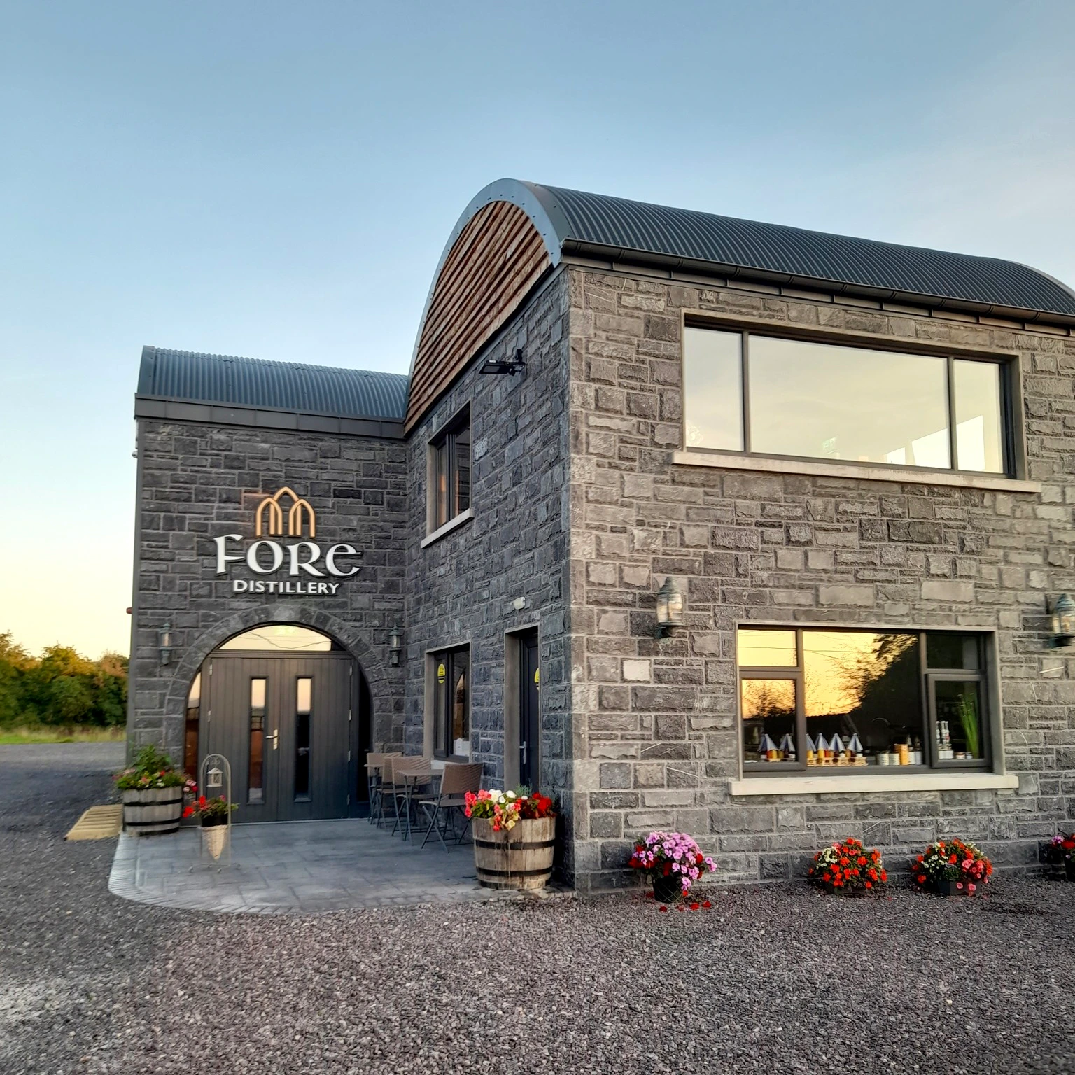 A stone building labeled "Fore Distillery" with large windows, outdoor seating, potted plants, and a gravel pathway.