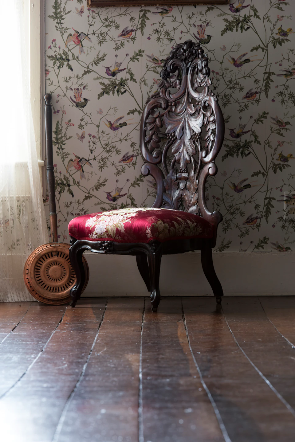 Ornate wooden chair with red floral upholstery against a bird-patterned wallpaper; a banjo and sheer curtain are visible nearby.
