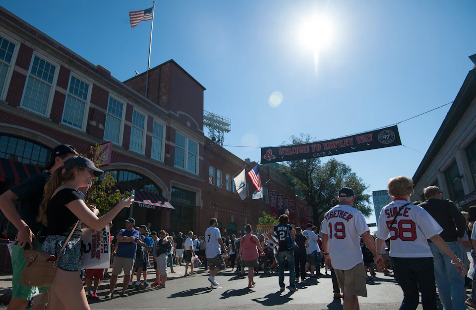 Crowd gathers outside a brick stadium under a banner that reads "Welcome to Yawkey Way" on a sunny day. People wear various sports jerseys.
