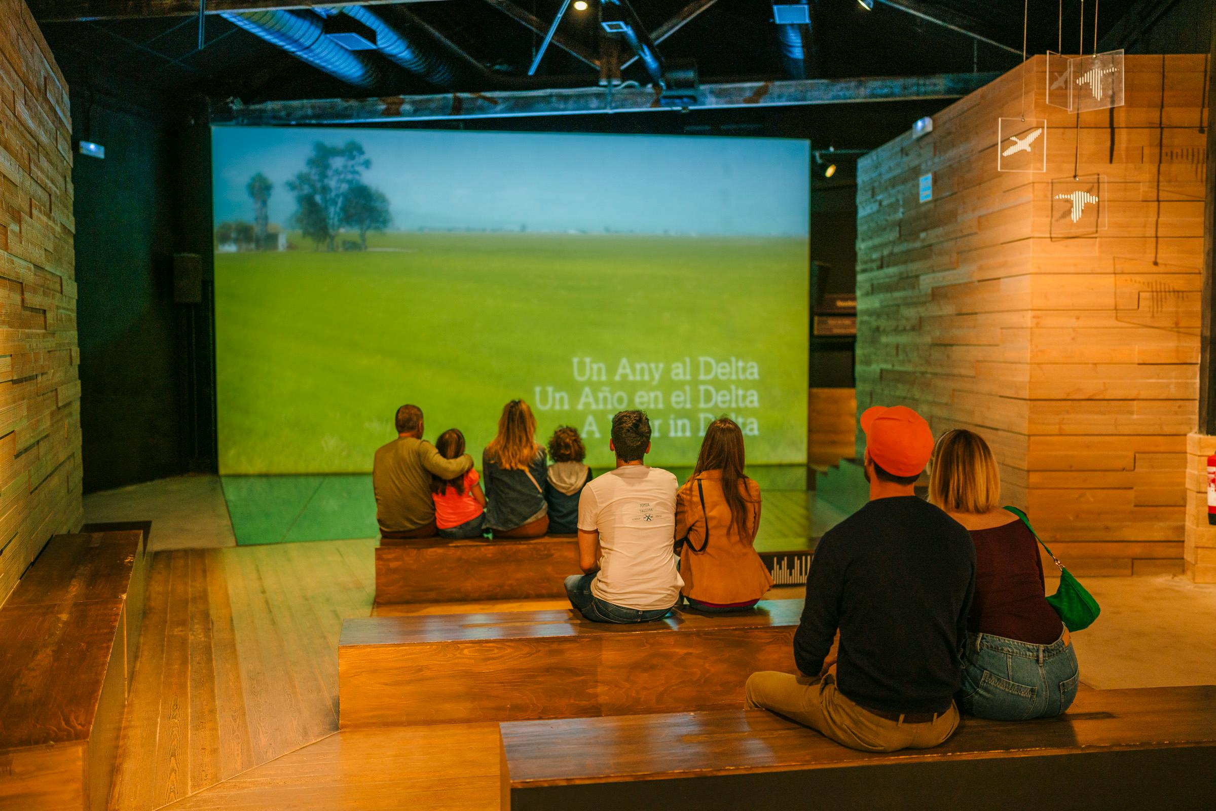 Lidé sedí na dřevěných schodech a sledují promítané video pole s textem „Un Año en el Delta“ ve více jazycích.