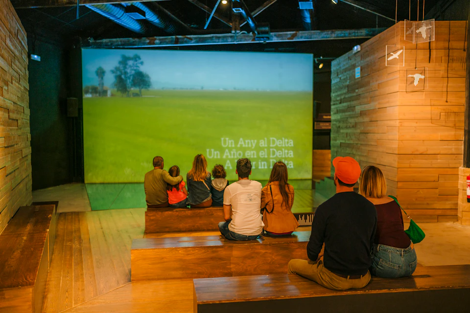 People sitting on wooden steps, watching a projected video of a field with the text "Un Año en el Delta" in multiple languages.
