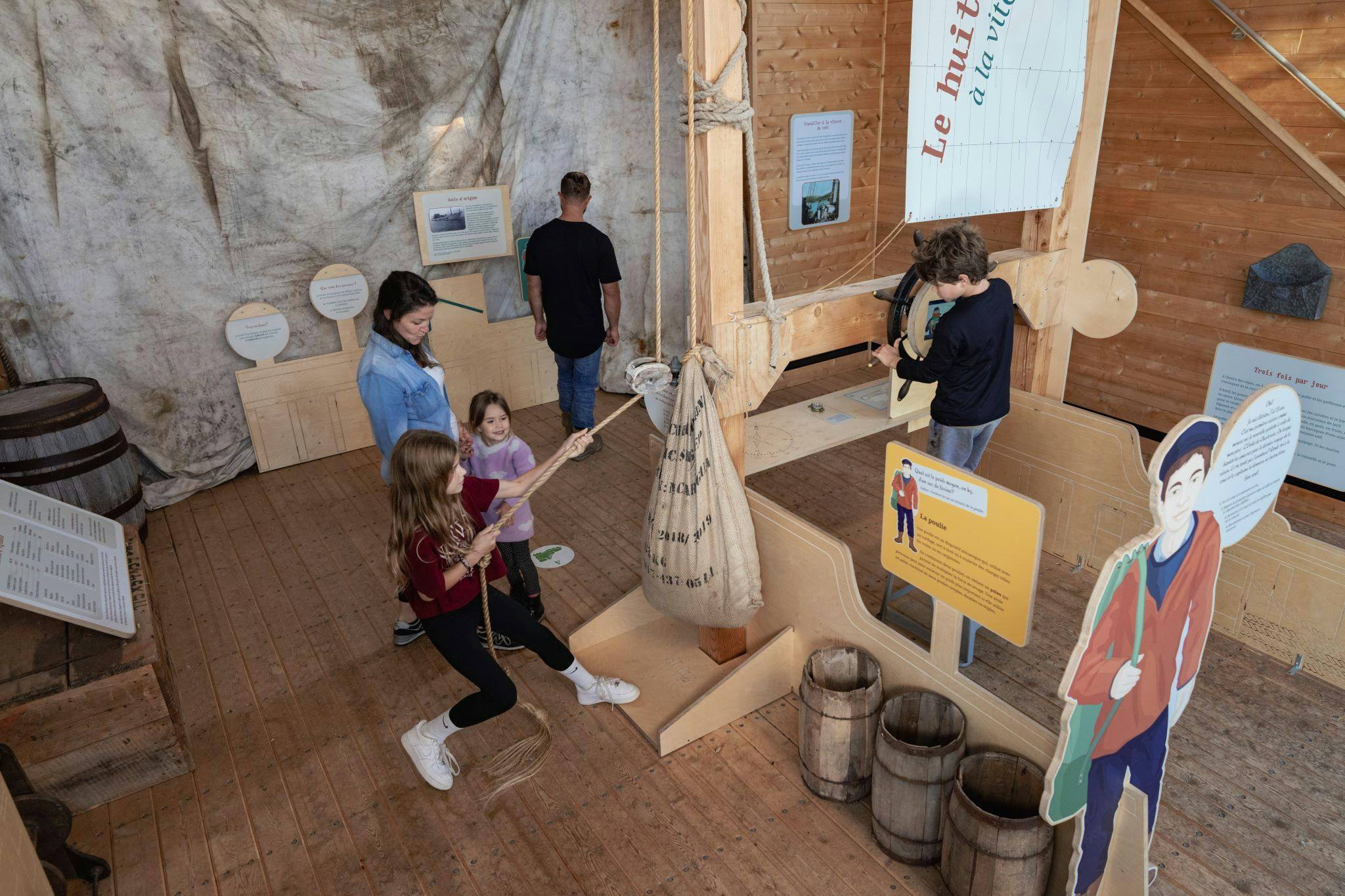 Børn og voksne inde i en trækonstruktion; to piger trækker i et reb, der er fastgjort til en sæk, mens to voksne observerer udstillinger.