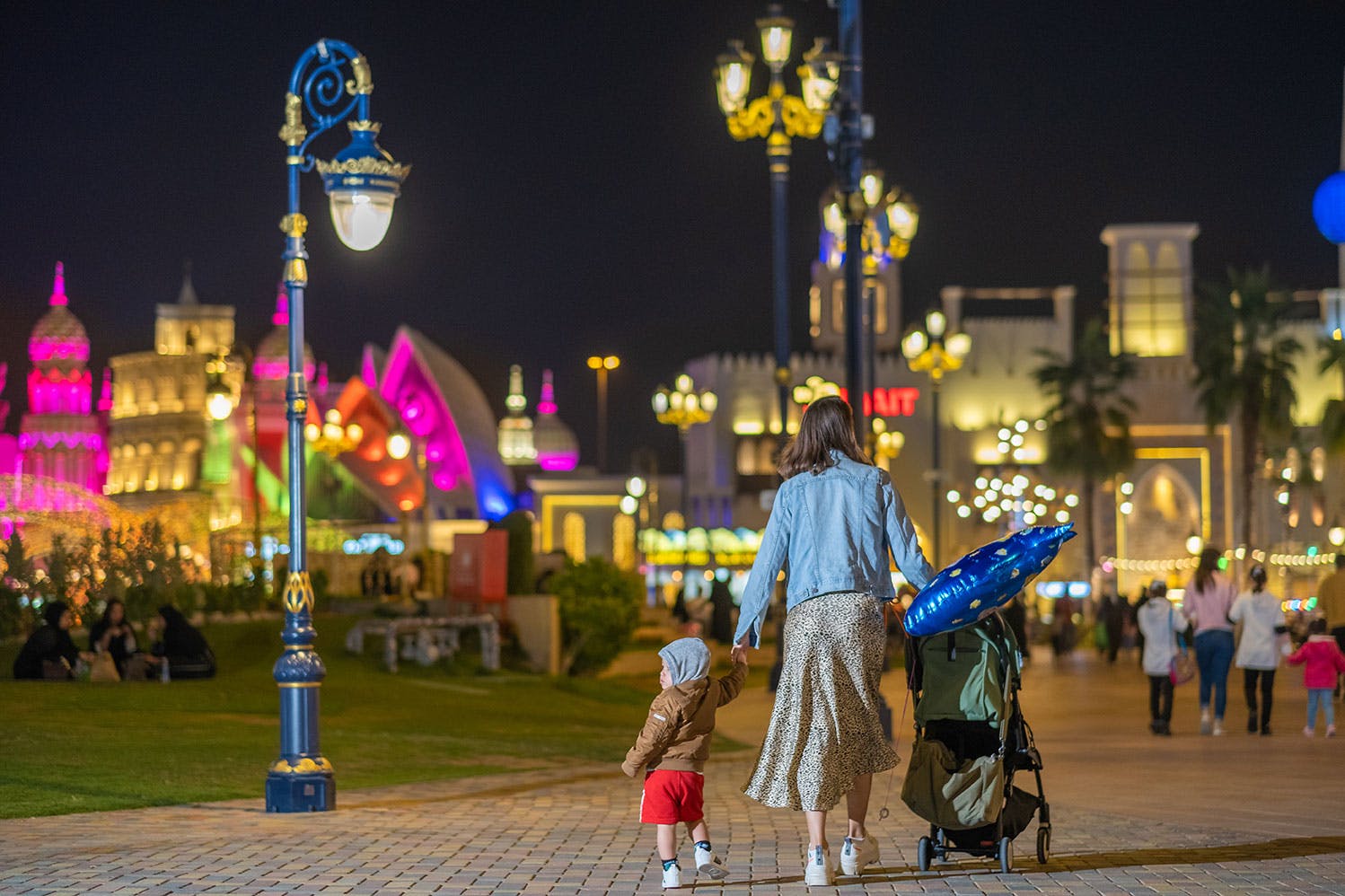 밤에는 가로등과 유모차가 있는 화려하고 밝은 야외 환경에서 한 여성과 어린이가 조명이 켜진 통로를 걷고 있습니다.