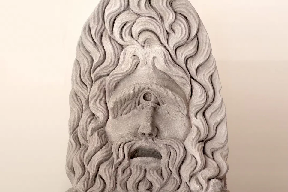 A stone sculpture of a bearded face with a single eye in the center of the forehead, framed by wavy hair.
