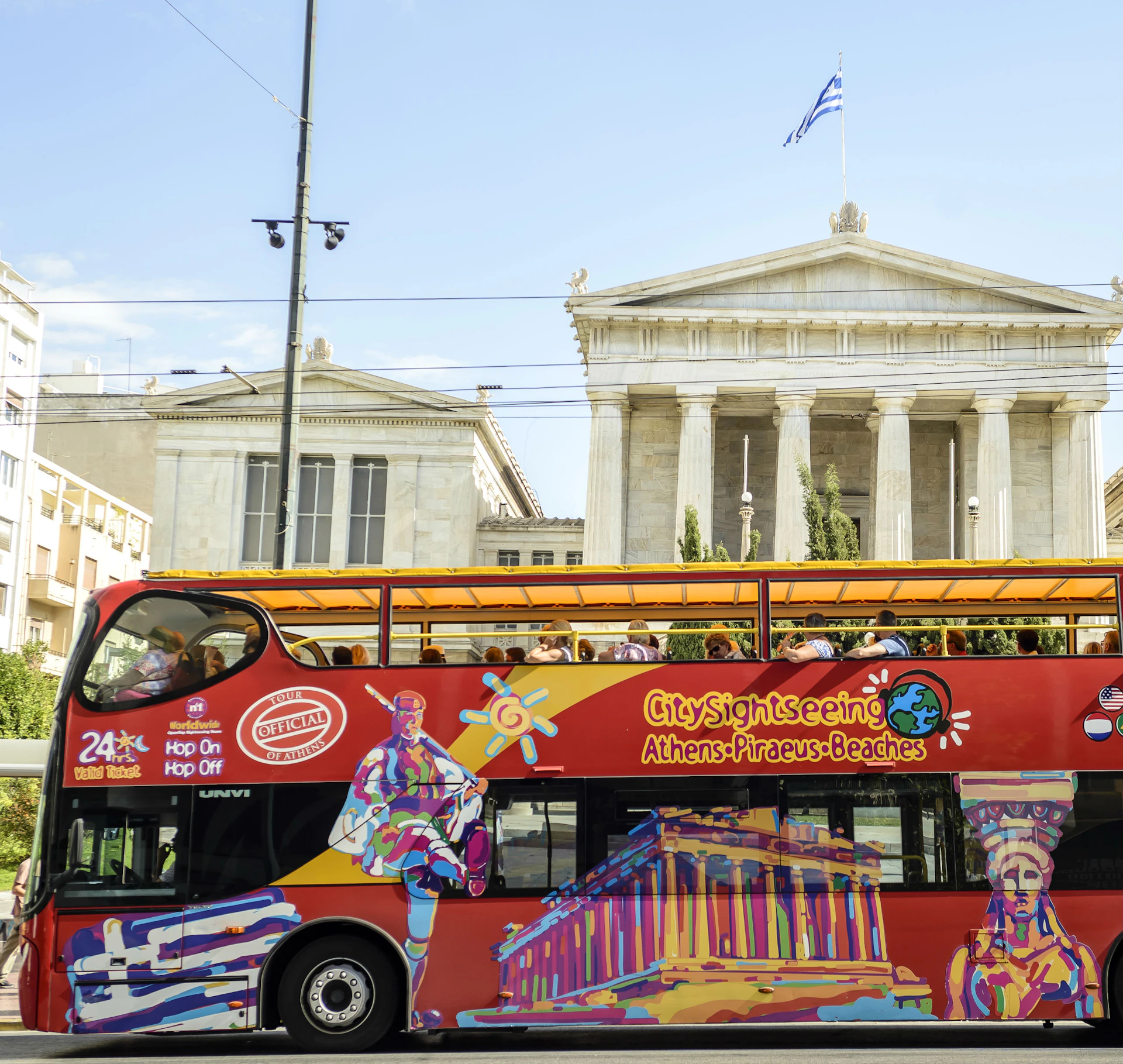 A vibrant, double-decker sightseeing bus with Athens landmarks painted on it passes in front of classical Greek architecture.
