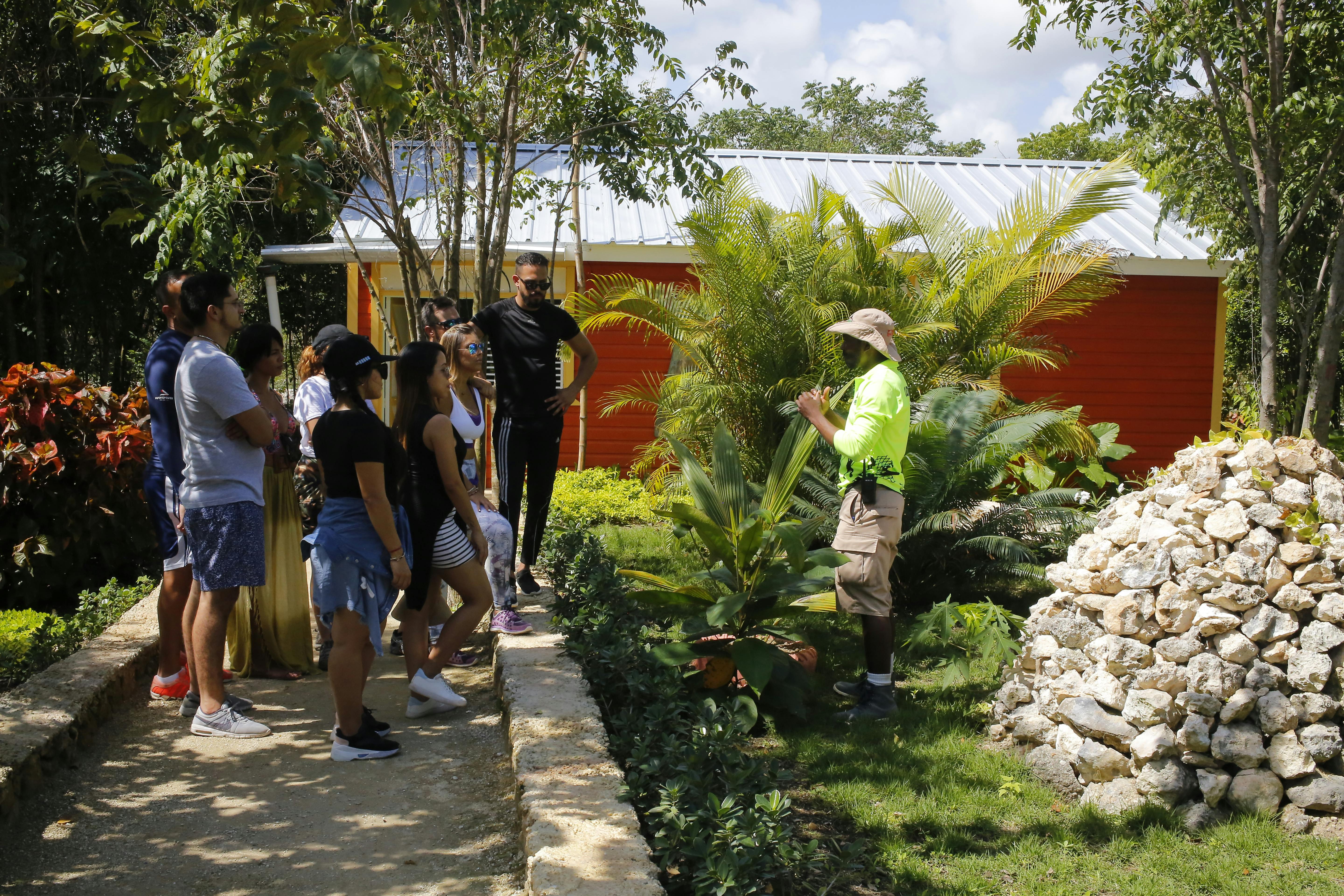 A group of people listens to a guide in a garden with greenery and a building with a tin roof and orange wall in the background.