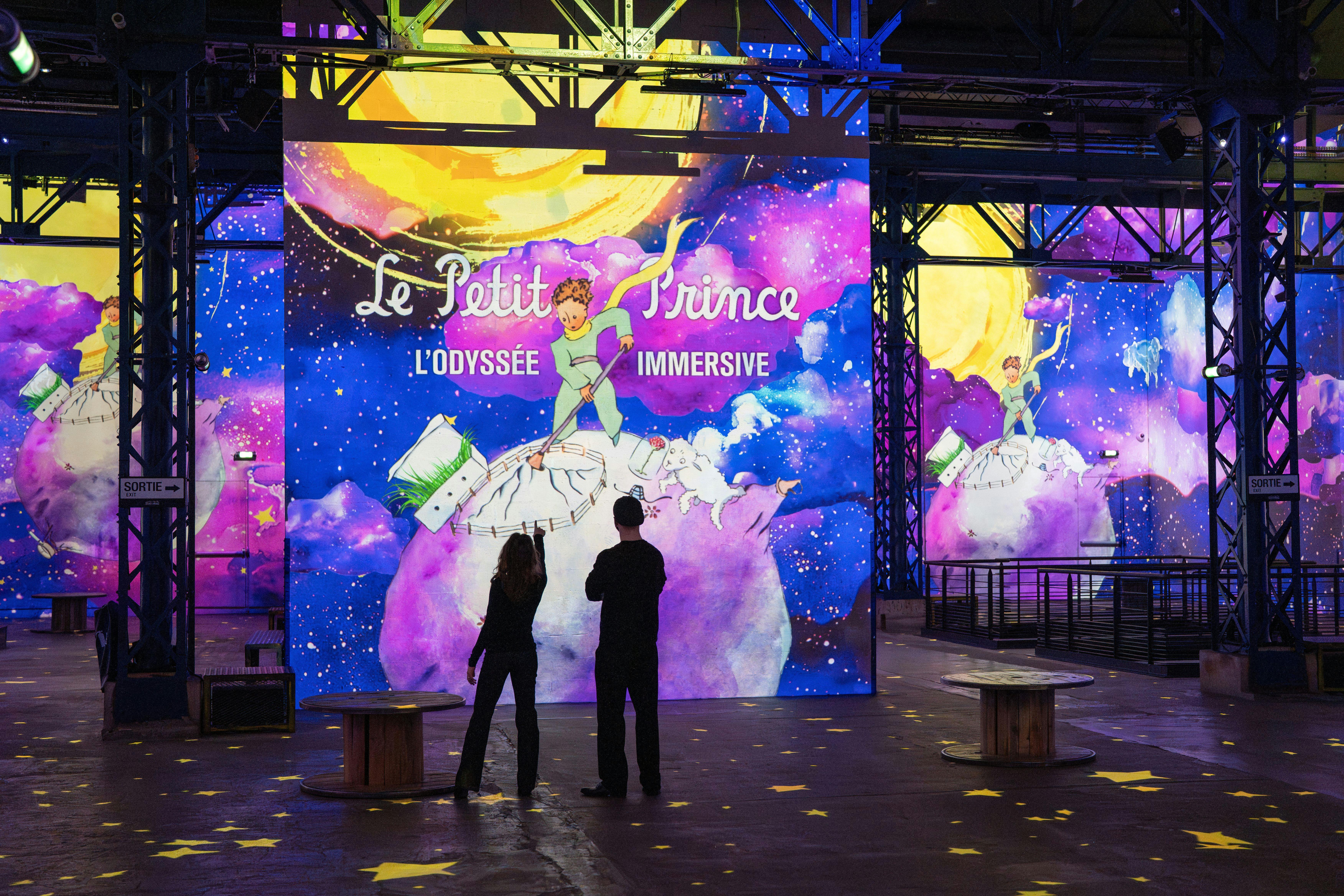 Two people stand in front of a colorful display featuring "Le Petit Prince: L'Odyssée Immersive" in a vibrant space.