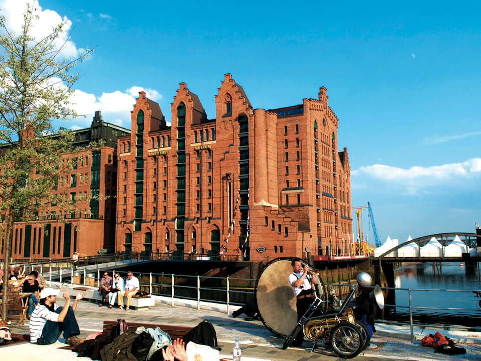 Brick building with arched windows by the water. In the foreground, a man plays a large drum near a railing. People are seated nearby.