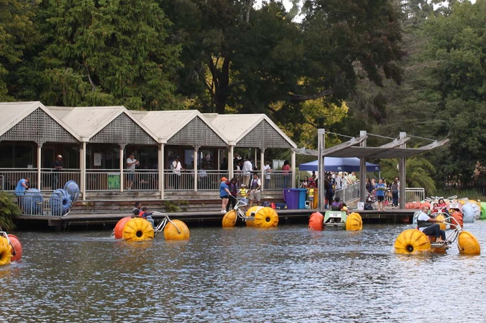 People enjoying a lakeside area with pedal boats shaped like oranges and a wooden building with a deck in the background.