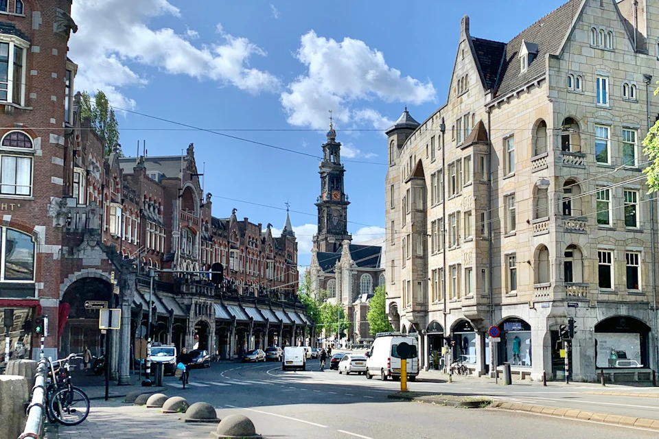 A street scene with historic brick buildings on both sides, a tall clock tower in the background, and vehicles on the road.