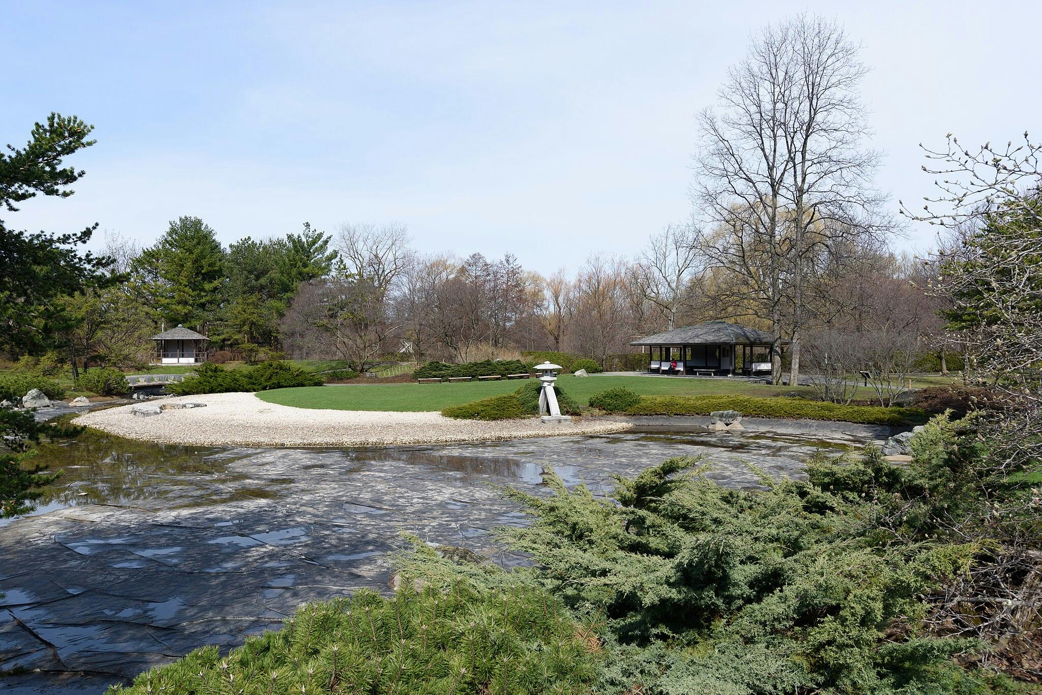 Ένας γαλήνιος κήπος με γκαζόν, χαλίκι, λιμνούλα, δύο κιόσκια και ένα πέτρινο φανάρι. Δέντρα και θάμνοι περιβάλλουν την περιοχή.