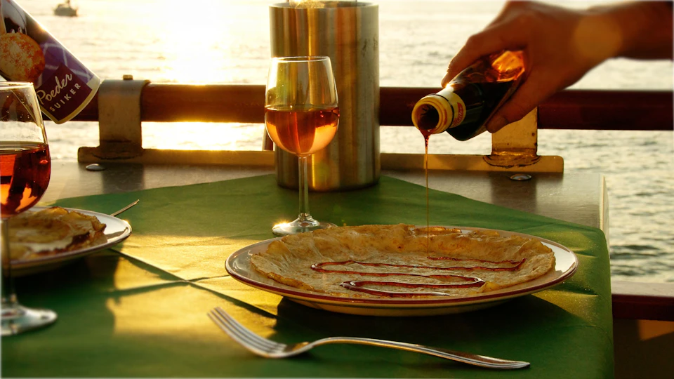 A person pours syrup onto a crepe on a plate next to a glass of wine, with a sunset view in the background.
