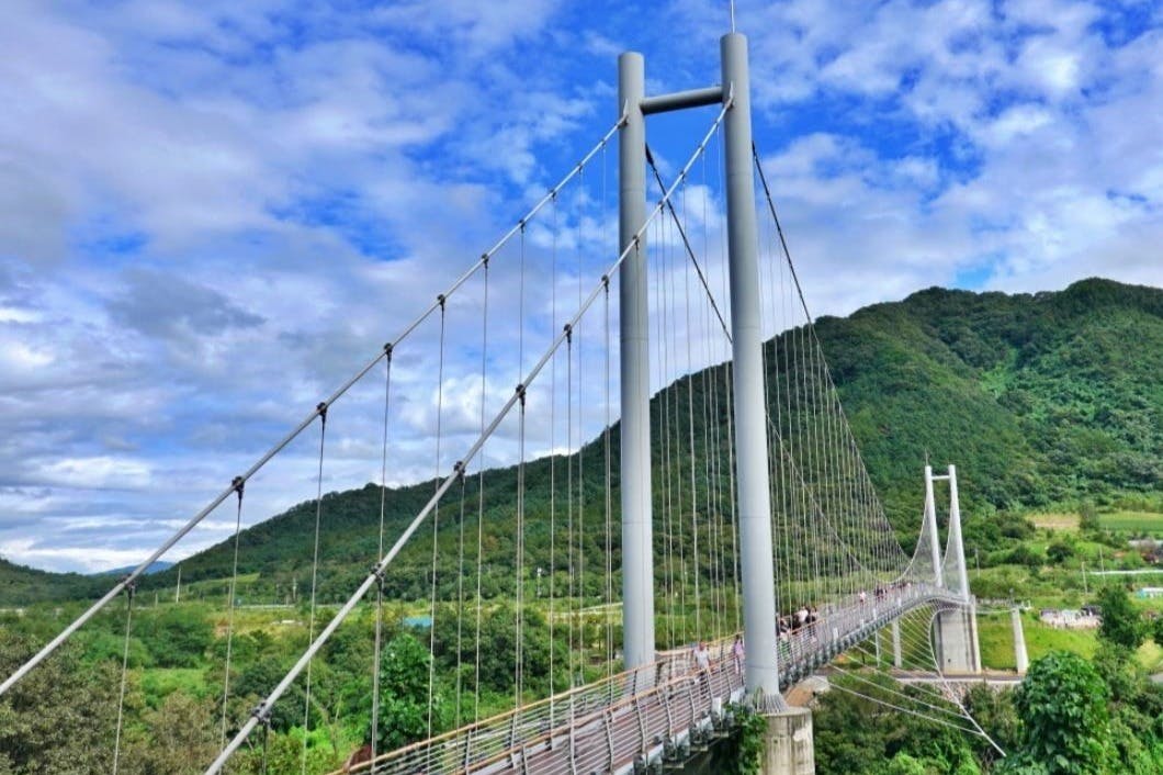 A suspension bridge spanning over a lush green valley, with a mountainous backdrop under a partly cloudy blue sky.
