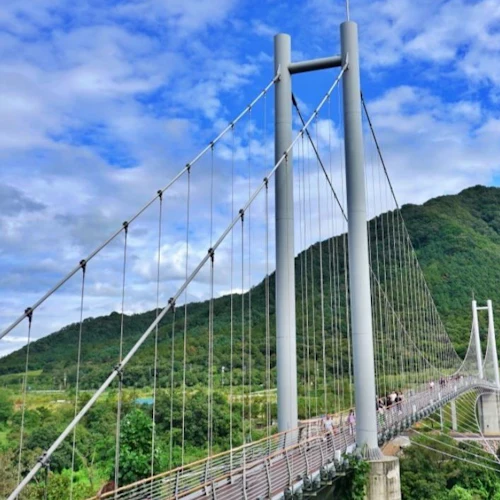 A suspension bridge spanning over a lush green valley, with a mountainous backdrop under a partly cloudy blue sky.