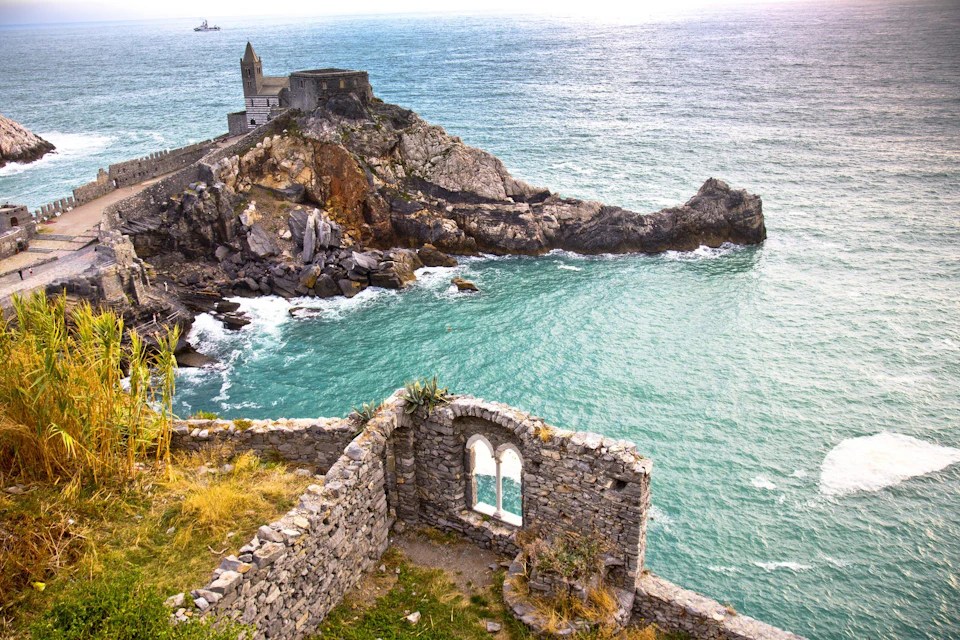 A coastal scene with a rocky outcrop featuring a small stone building and an ancient stone arch in the foreground. Turquoise sea below.