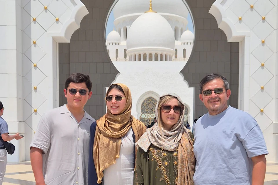 Three people in sunglasses pose in front of a white mosque with a domed roof and ornate architectural details.