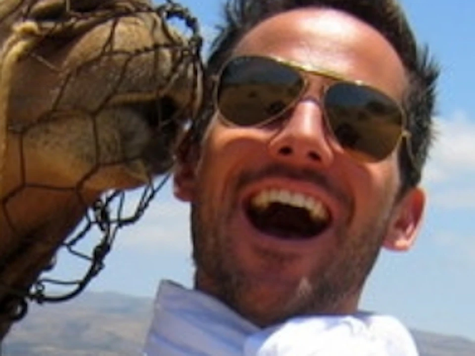 A man laughs while a nearby camel nuzzles his face. A woman smiles in the background, seated on a green cart.
