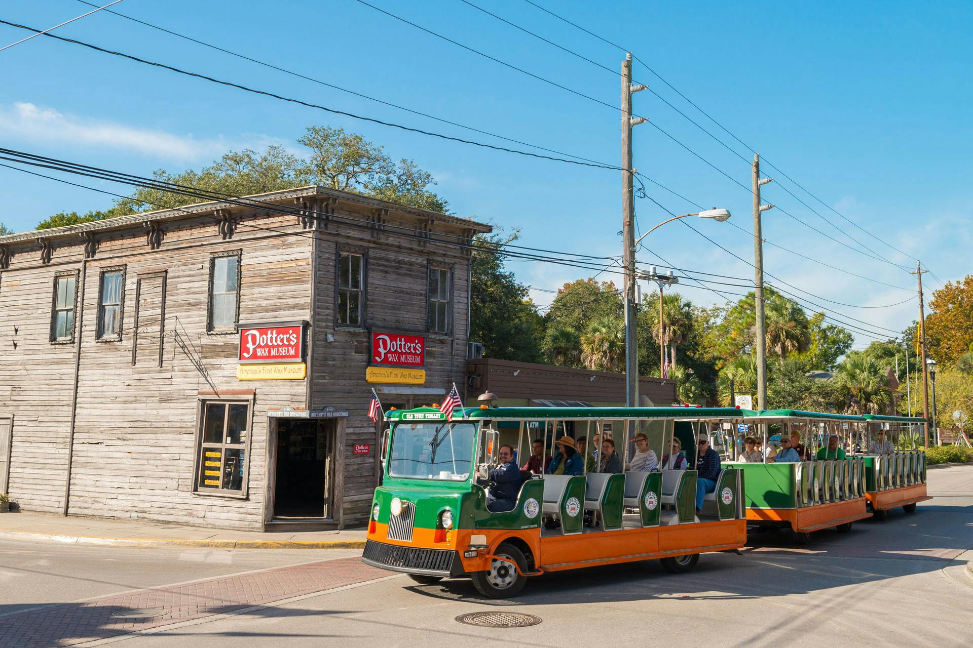 A green and orange tourist tram passes a rustic building labeled "Potter's Wax Museum" under a clear blue sky.