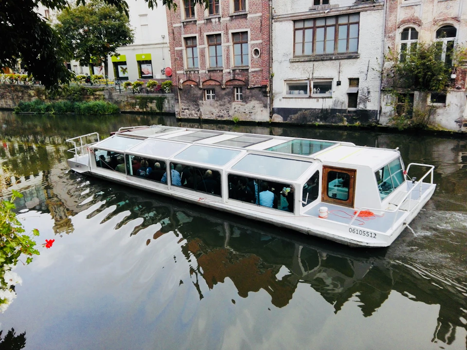 White canal boat with glass roof cruising on a river, with brick and stone buildings in the background.
