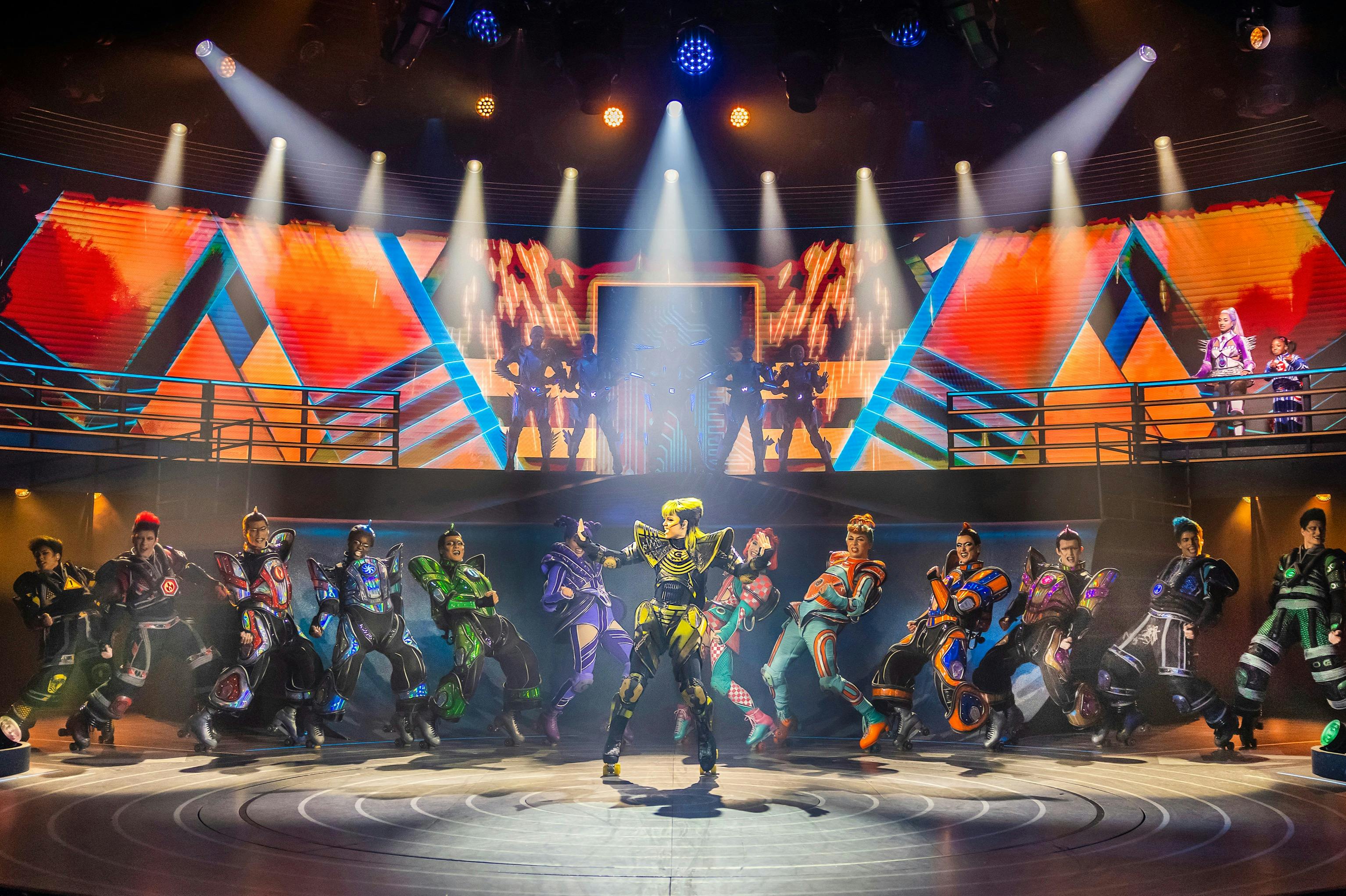 身着色彩缤纷的未来派服装的表演者在舞台上跳舞，舞台上有充满活力的数字屏幕和聚光灯效果。