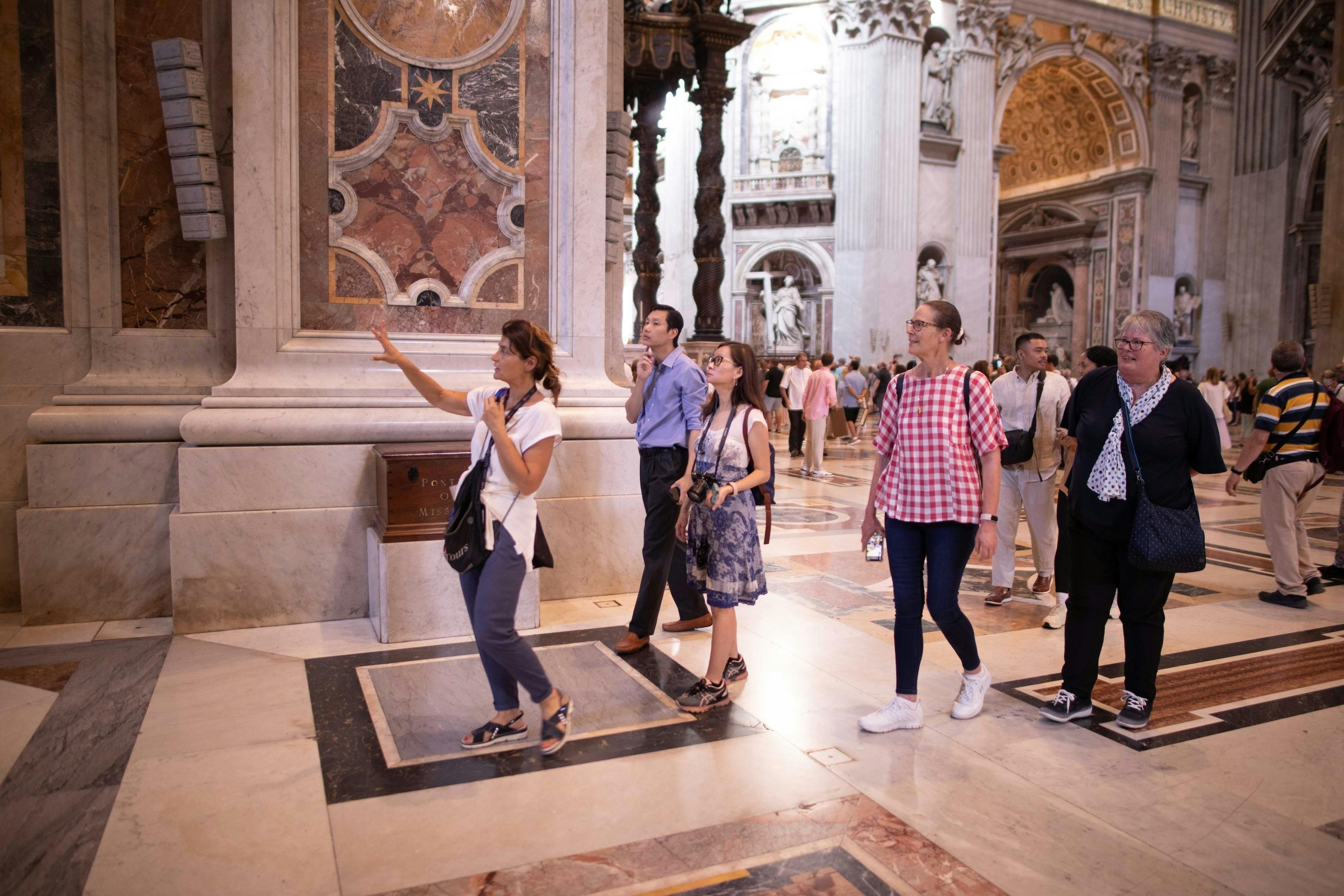 Un gruppo di turisti che ammira delle opere d'arte all'interno di un edificio maestoso e riccamente decorato, con pavimenti in marmo e pareti ornate.
