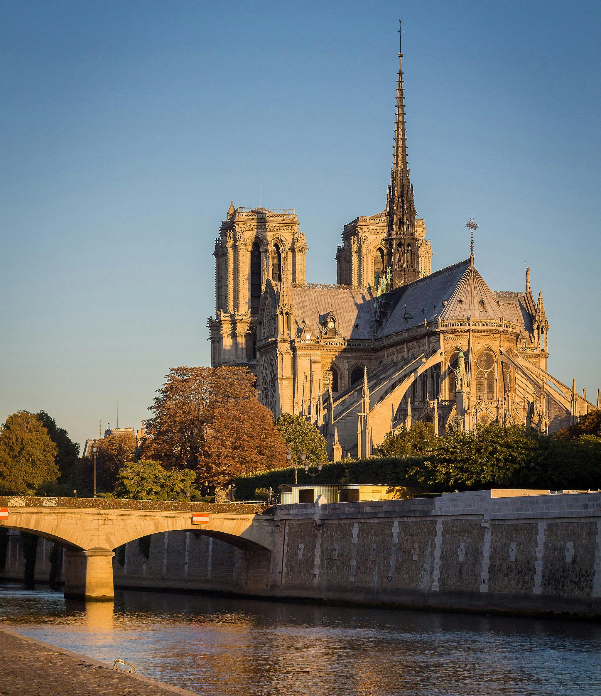 Cathedral with prominent spire and two towers, beside a river, with a stone bridge and lush trees in the foreground.