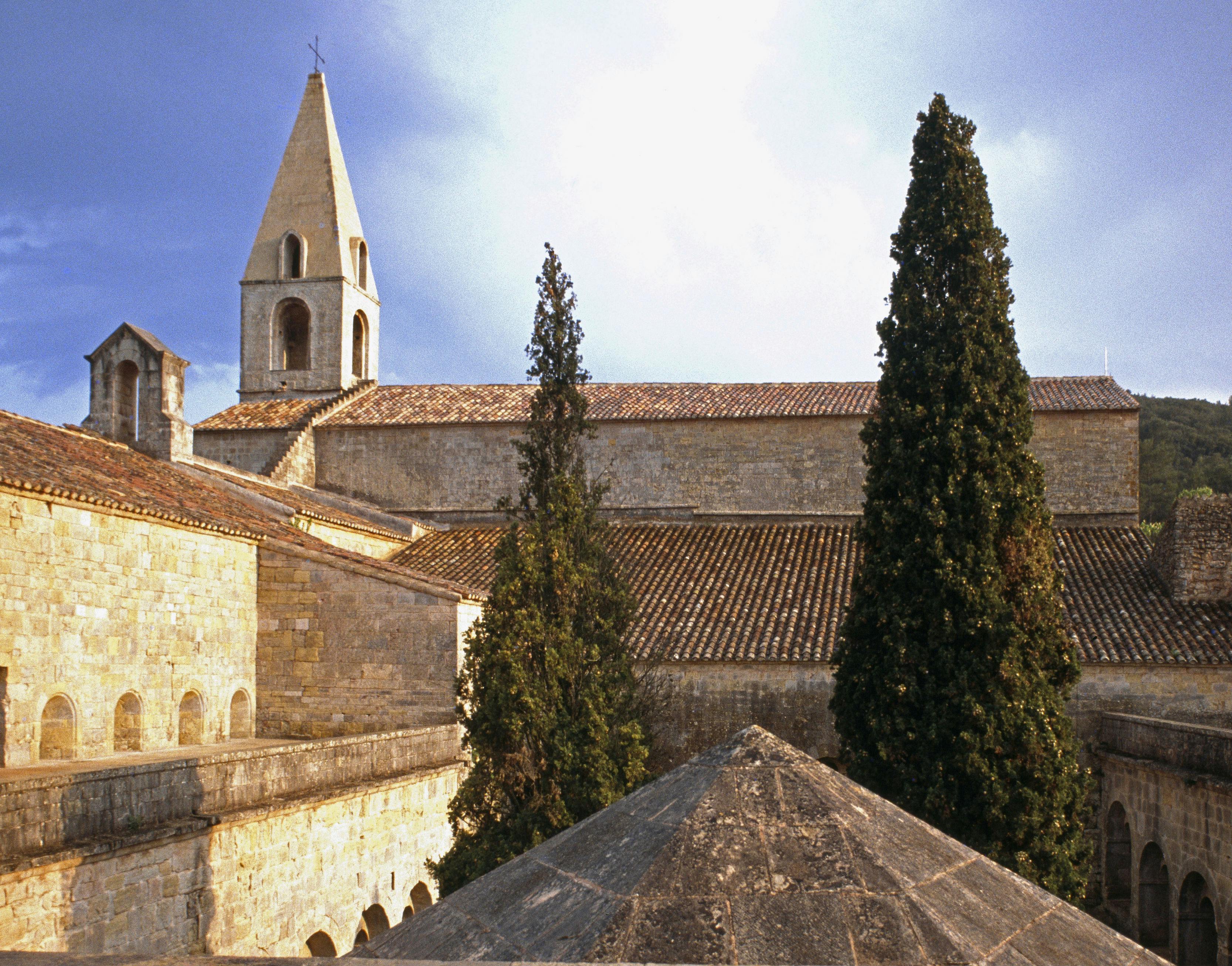 Πέτρινη εκκλησία με μυτερό καμπαναριό, ψηλά κυπαρίσσια και κεραμοσκεπή κάτω από τον γαλάζιο ουρανό.