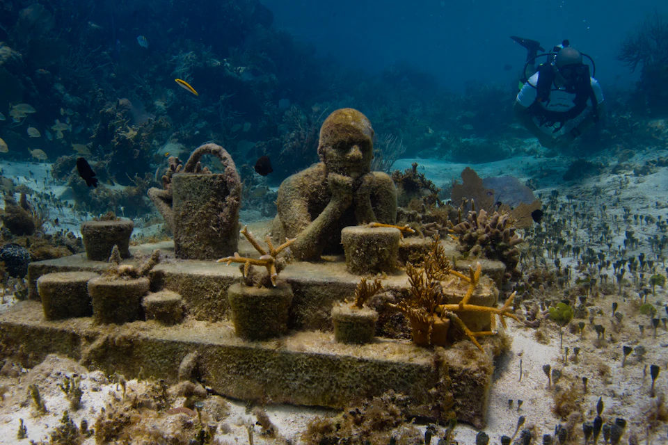Underwater scene with a coral-covered statue of a person leaning on their hands, surrounded by marine life and a diver in the background.