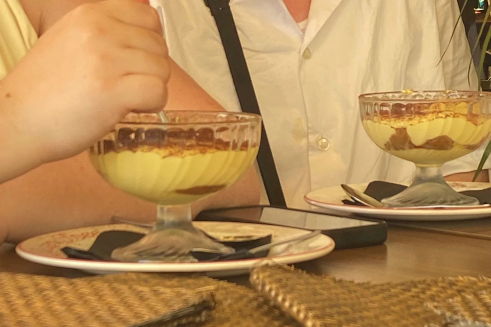 Two people are seated, eating creamy desserts from glass bowls. A smartphone and woven placemats are on the table.