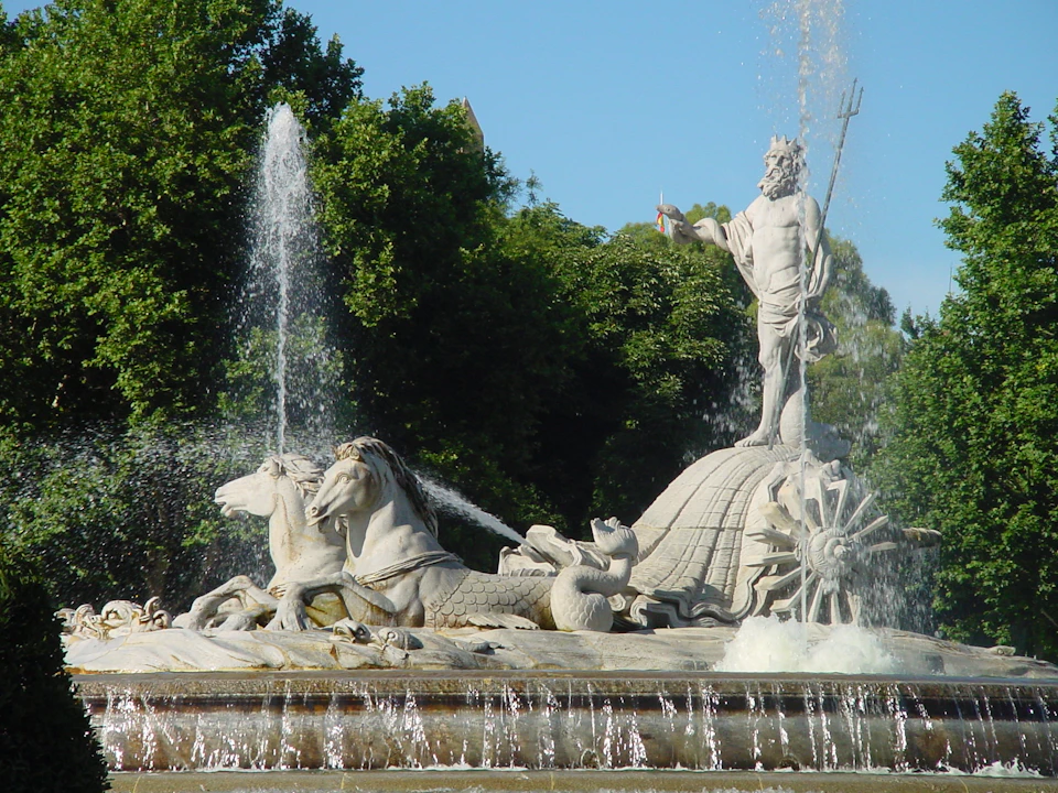 Statue of Neptune holding a trident, standing on a chariot with horses and water spraying fountains in a garden setting.