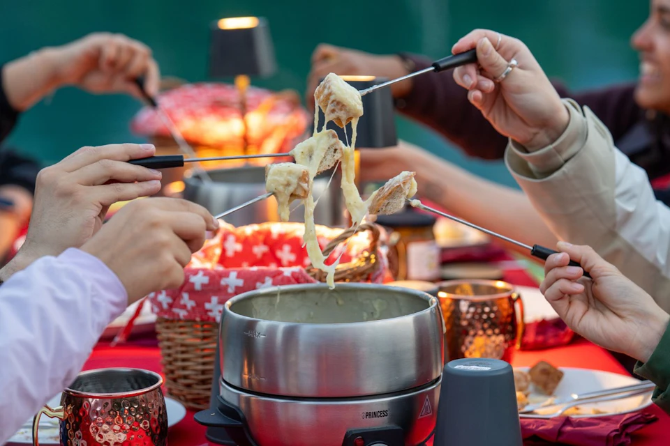 People holding fondue forks with melted cheese above a fondue pot on a table with red decorations and baskets.