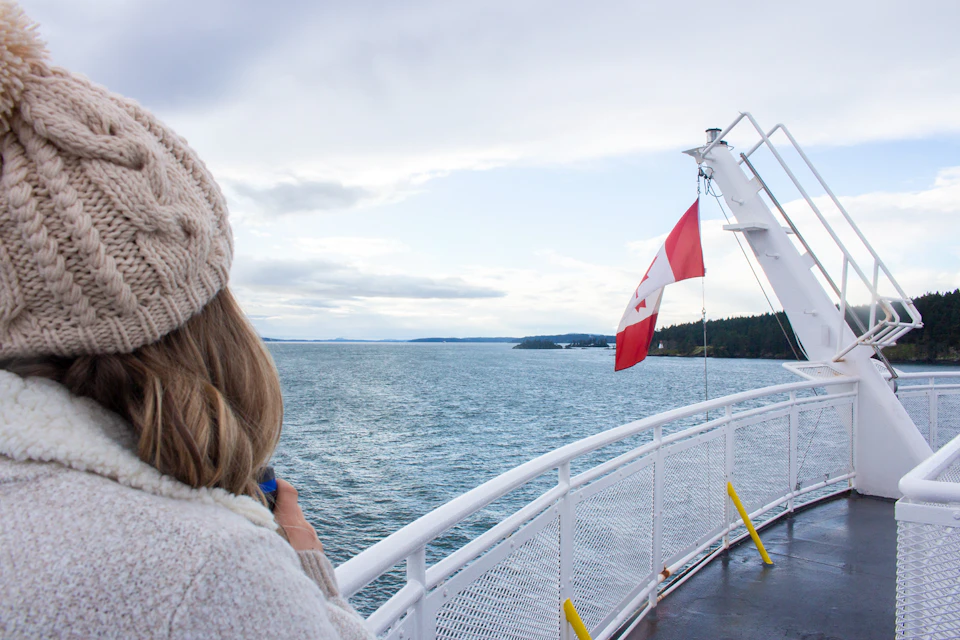 Person in a knit hat looking out over the water from a ferry with a Canadian flag, distant land visible.