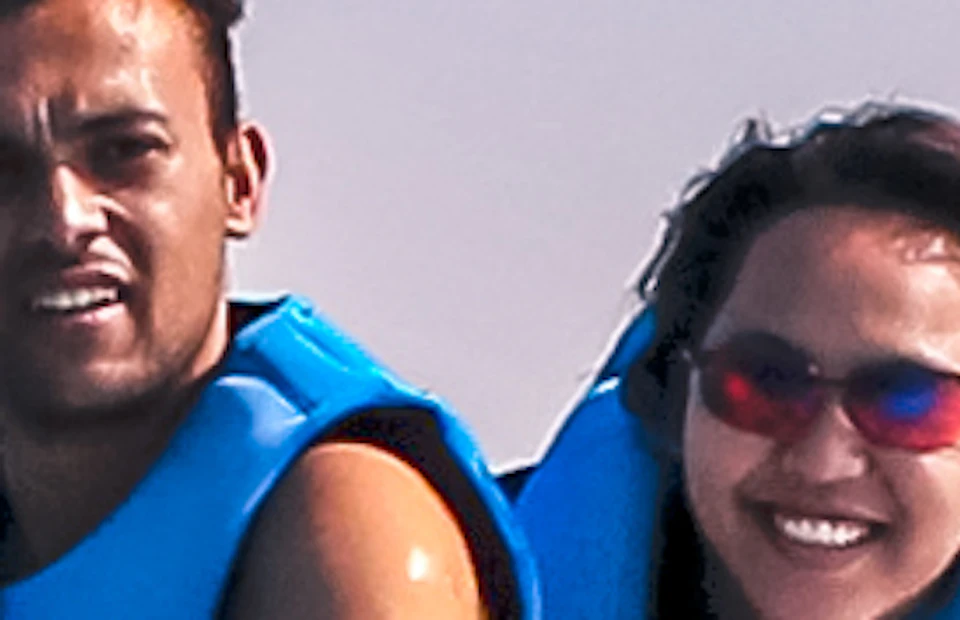 Two people riding a jet ski on open water, with a large, unique building and construction cranes visible in the background under a clear sky.