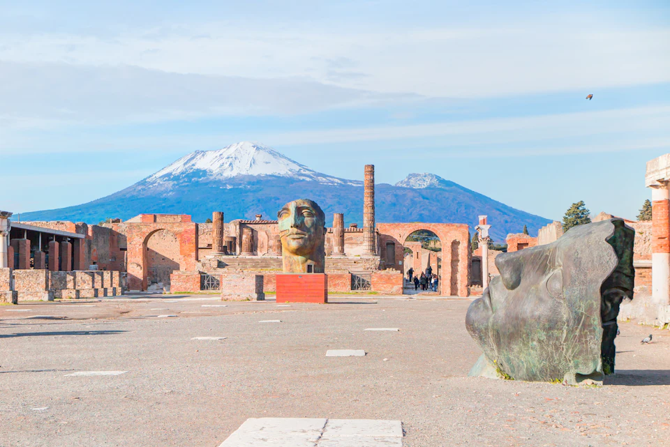 Ancient ruins with large bronze head sculptures in foreground. Snow-capped mountains in the background under a clear sky.