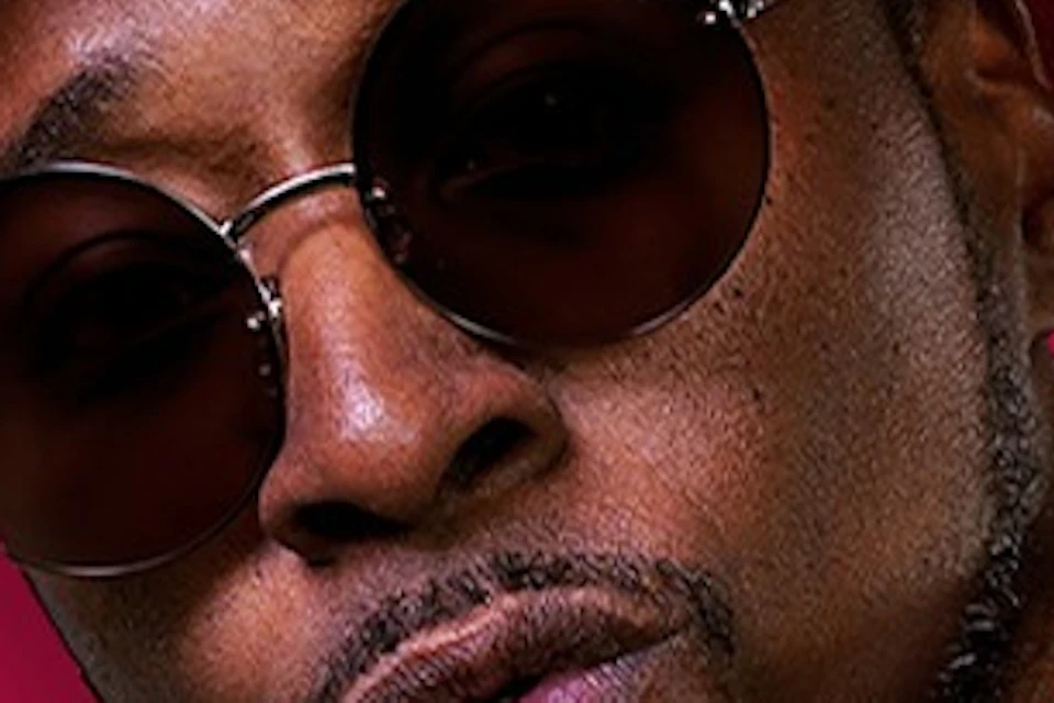 A person poses wearing a sequined jacket, a red shirt, sunglasses, and a hat, with a magenta background.