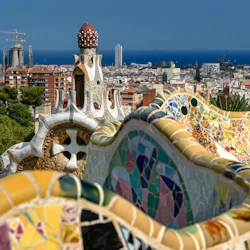Färgglada mosaikplattor och nyckfull arkitektur i Park Güell med utsikt över Barcelonas stadsbild och det avlägsna blå havet under en klar himmel.