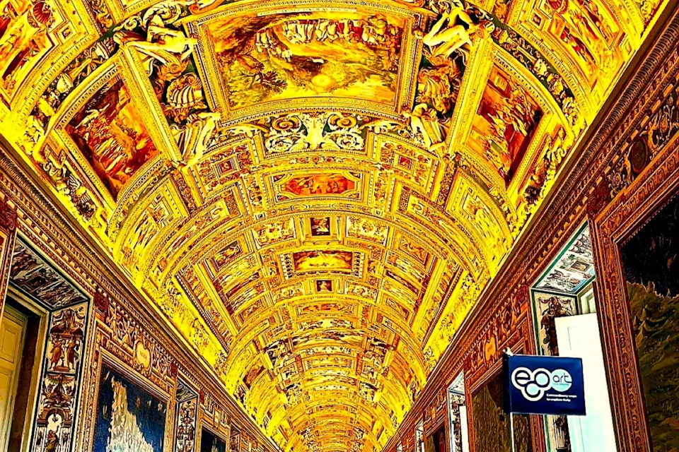 An ornately decorated vaulted ceiling with detailed paintings and gold accents in a richly adorned corridor.