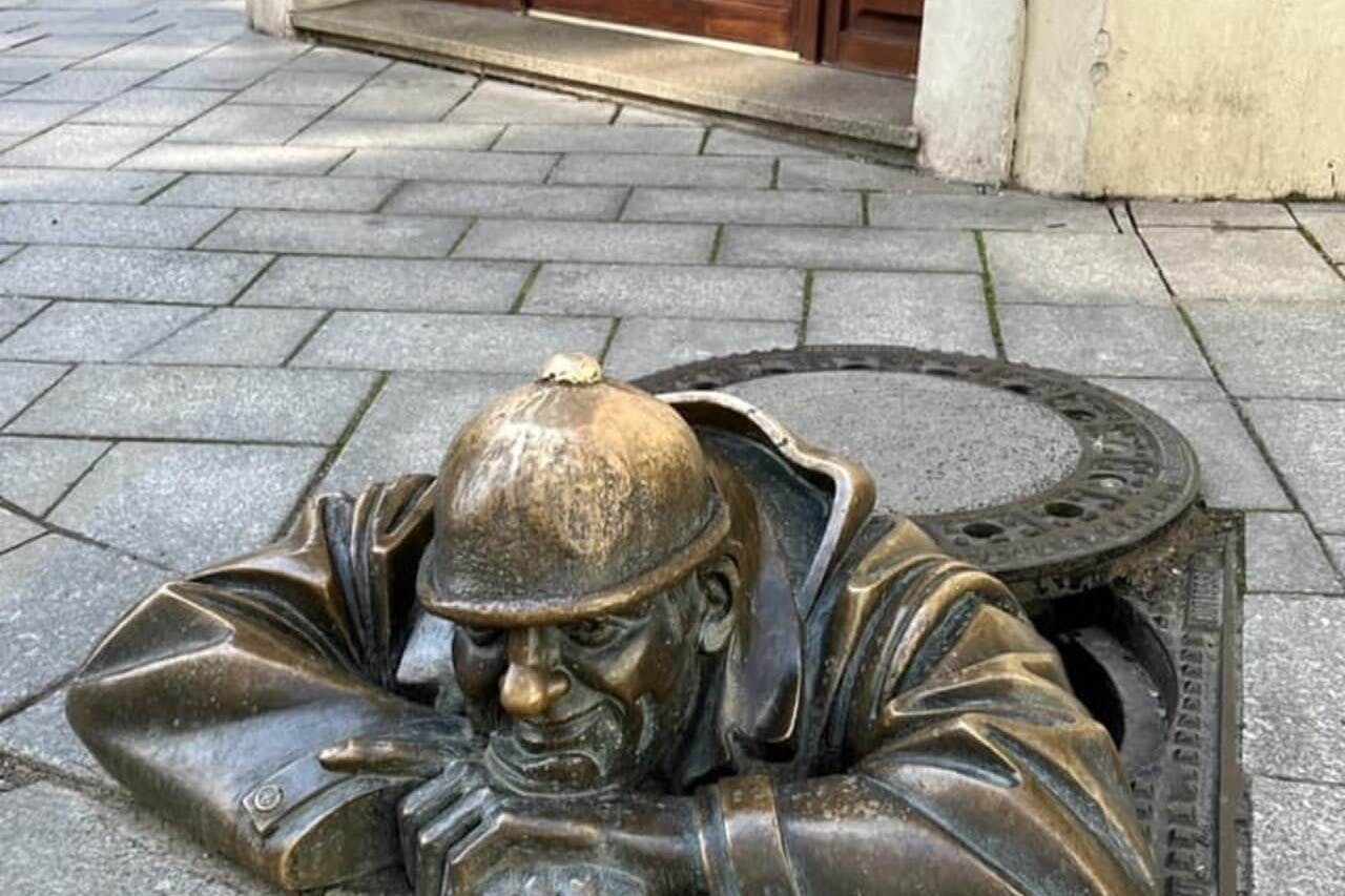 在建筑入口附近的铺面人行道上，一名男子从人孔中窥视的青铜雕像。