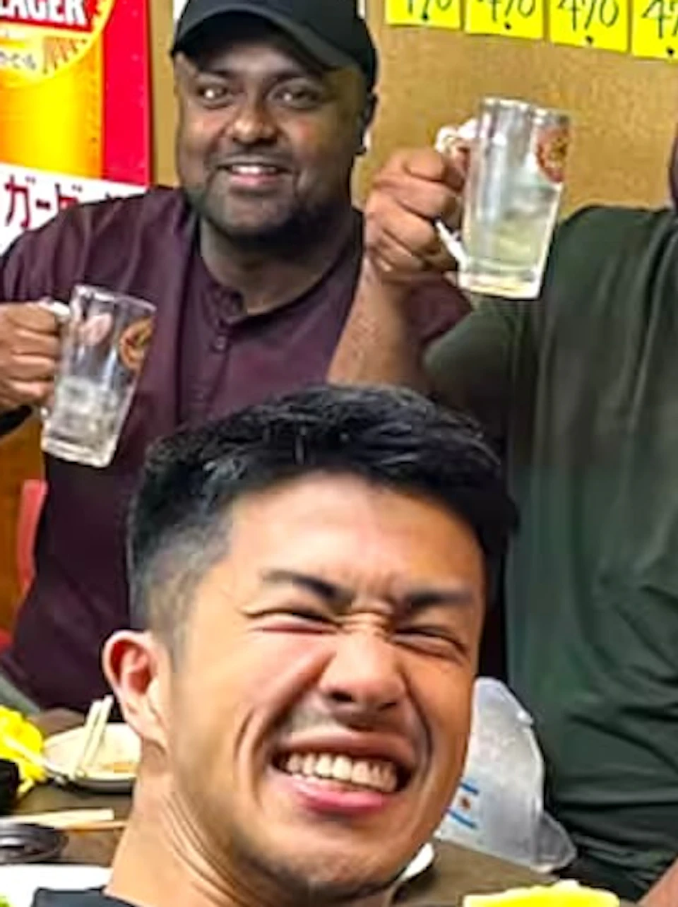 A group of people sitting around a table in a restaurant, smiling and holding bottles and glasses, with food on the table.